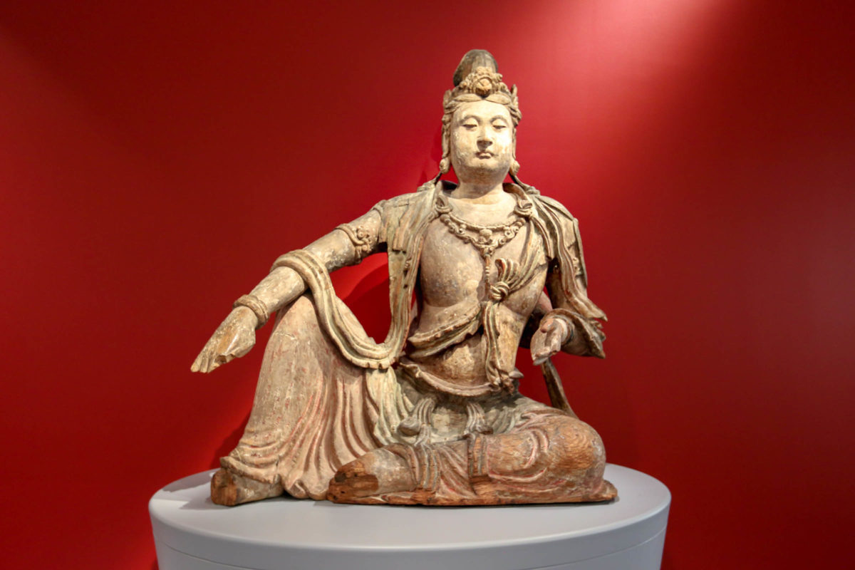 A wooden carving on Guanyin on a gray pedestal in front of a red wall.
