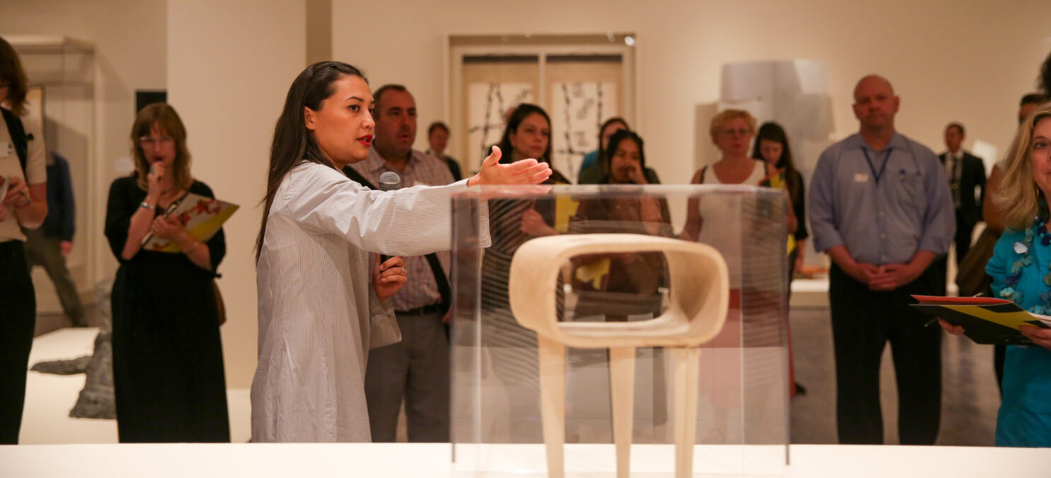 A curator gestures toward a contemporary sculpture in front of a crowd of visitors