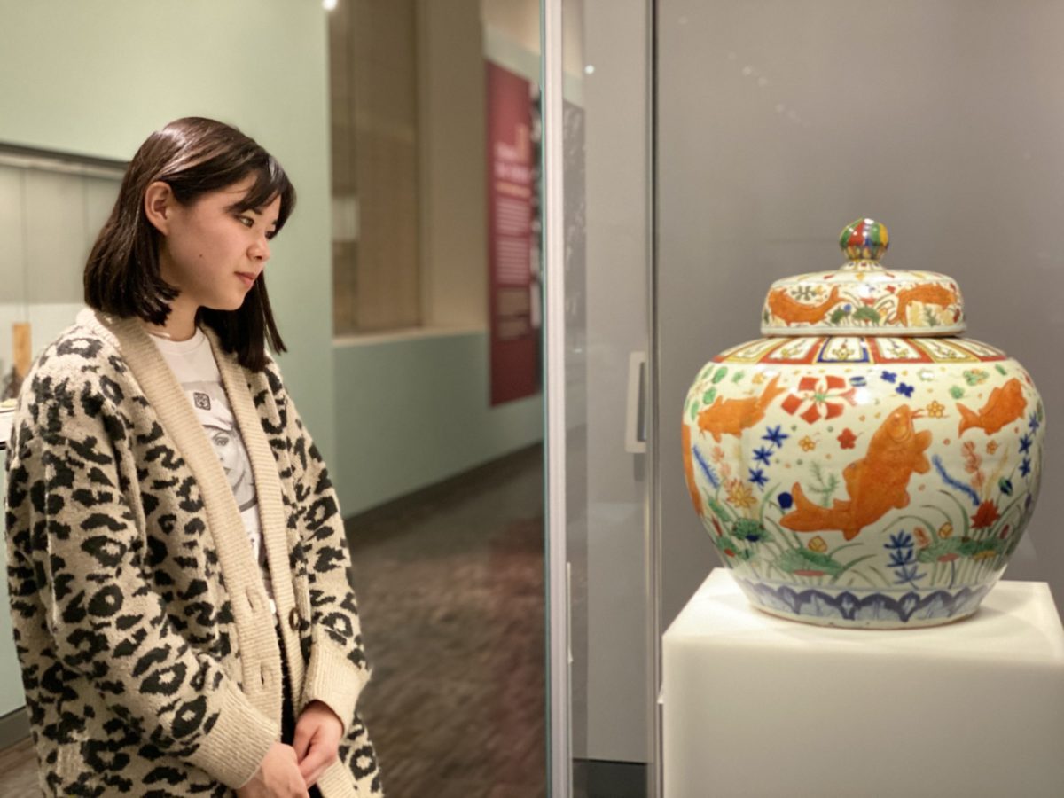 A woman in a leopard print sweater looks at a jar painted with fish and flower designs.