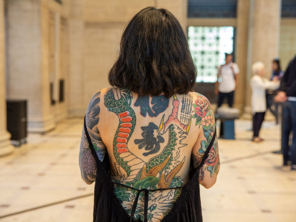 A woman with short dark hair and an open back dress shows off a dragon tattoo that covers her back.